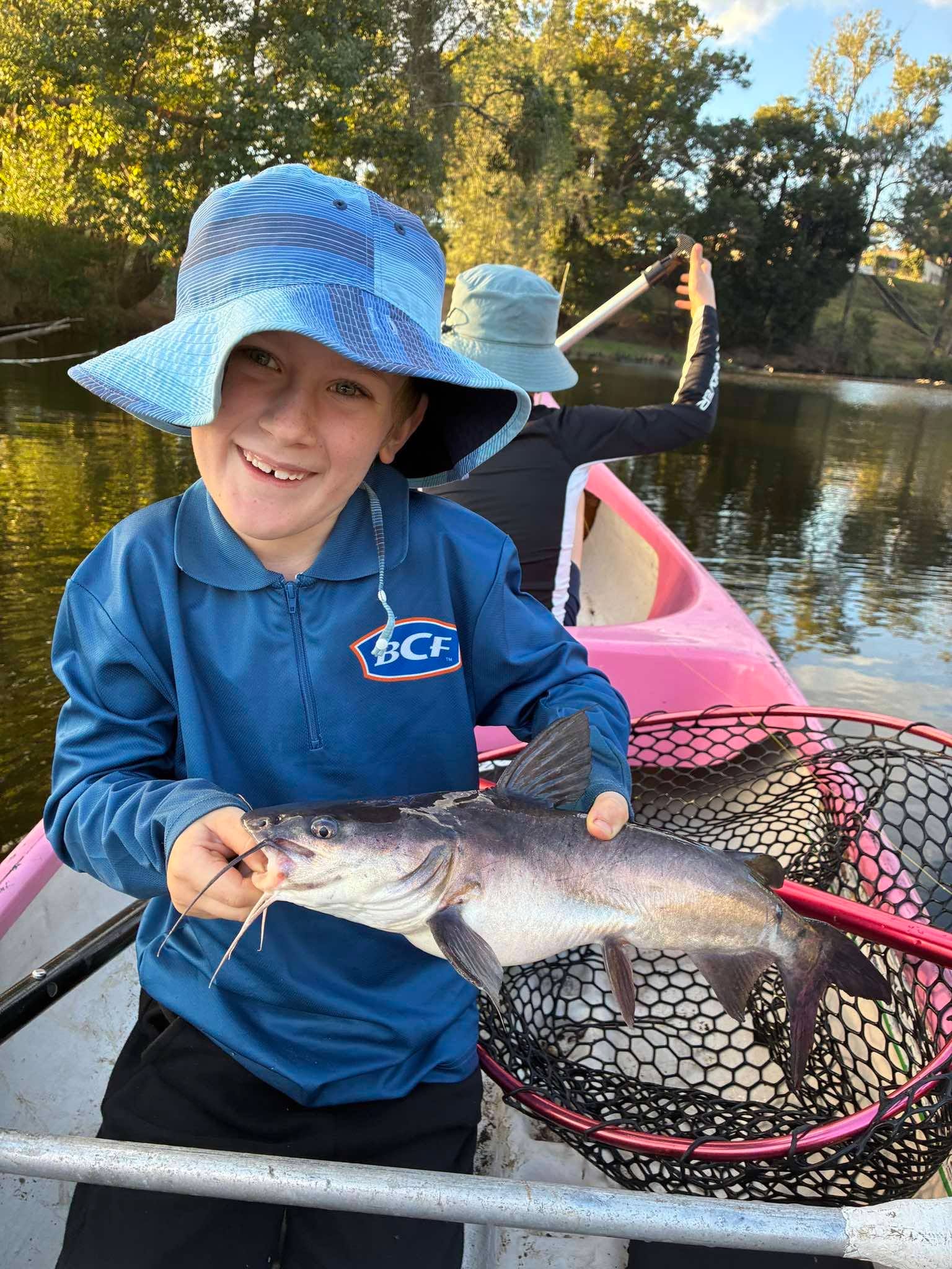 A young camper holding a fish in a canoe on the tree-lined creek